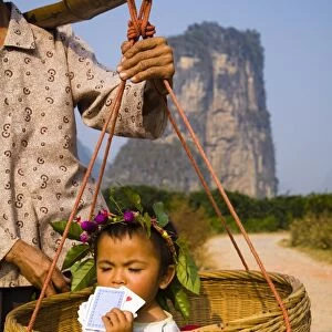 China, Guangxi Zhuang Autonomous Region, Yangshuo County. Young chinese child being carried in an hand woven basket along a rural road running through an agriculture landscape dominated by the Karst peaks of