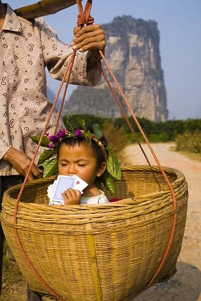 China, Guangxi Zhuang Autonomous Region, Yangshuo County. Young chinese child being carried in an hand woven basket along a rural road running through an agriculture landscape dominated by the Karst peaks of