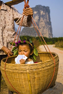 China, Guangxi Zhuang Autonomous Region, Yangshuo County. Young chinese child being carried in an hand woven basket along a rural road running through an agriculture landscape dominated by the Karst peaks of